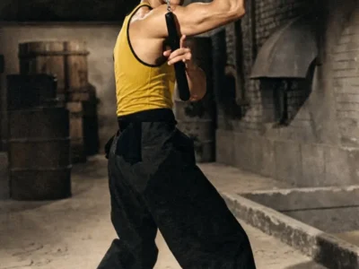 An archival-style campaign image for the LỰU ĐẠN x Bruce Lee collaboration features Bruce Lee posed mid-combat with nunchaku in an industrial alleyway setting. Wearing his iconic yellow tank top and black trousers, Lee’s sharply defined stance and intense expression capture the physical precision and cinematic tension that made him legendary. Bold yellow “LỰU ĐẠN” typography stretches across the foreground, reinforcing the fusion of martial arts mythology, Asian cultural defiance, and contemporary luxury streetwear rebellion that defines the capsule collection