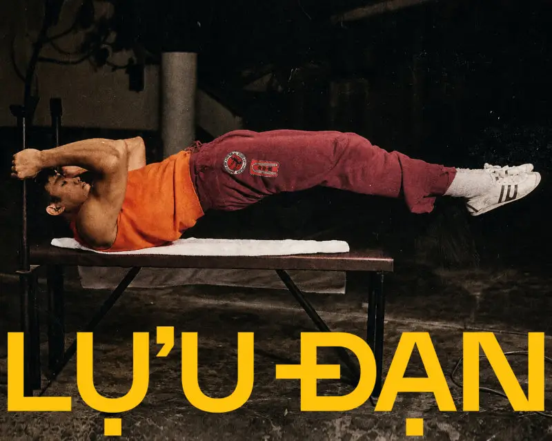 An archival-style training image from the LỰU ĐẠN x Bruce Lee collaboration captures Bruce Lee performing an intense core exercise while suspended horizontally on a workout bench. Dressed in a burnt orange tank top, burgundy training pants, and classic white sneakers, Lee’s disciplined physicality and athletic precision become the focal point within the dimly lit gym setting. Oversized yellow “LỰU ĐẠN” typography stretches across the foreground, reinforcing the capsule’s themes of endurance, masculine mythology, and explosive cultural defiance rooted in Bruce Lee’s enduring legacy