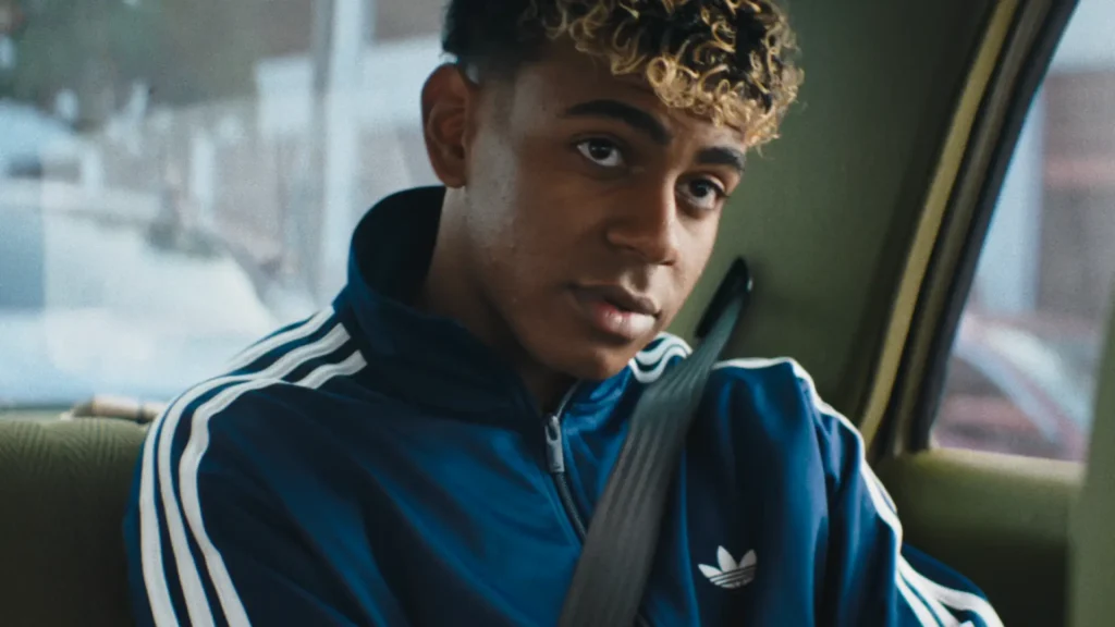A young man sits in the back seat of a car wearing a royal blue adidas track jacket with classic white three-stripe detailing and a matching Trefoil logo on the chest. A seatbelt crosses over the sporty silhouette as soft daylight filters through the windows, illuminating his curly highlighted hair and focused expression. The muted car interior and blurred cityscape outside create an intimate cinematic atmosphere, blending contemporary football culture with retro-inspired adidas styling and day urban realism