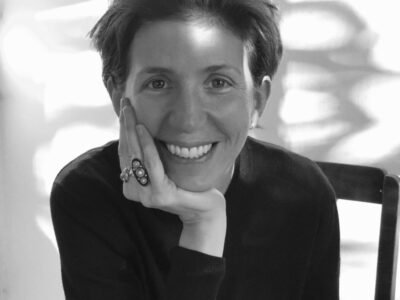 Black-and-white portrait of Ludivine Poiblanc seated indoors beside a chair, smiling warmly toward the camera with her hand resting against her face. She wears a minimalist dark knit top and a bold statement ring, while soft natural light casts abstract window shadows across the background, reflecting the understated elegance associated with her editorial and fashion direction work