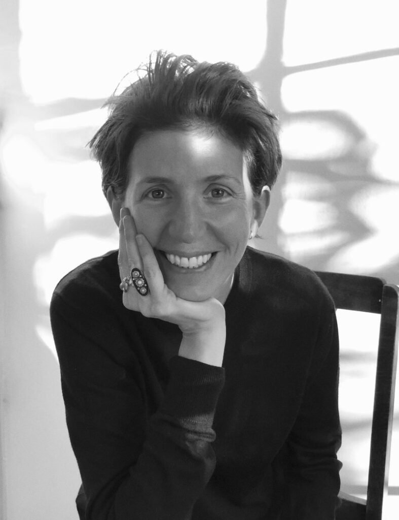 Black-and-white portrait of Ludivine Poiblanc seated indoors beside a chair, smiling warmly toward the camera with her hand resting against her face. She wears a minimalist dark knit top and a bold statement ring, while soft natural light casts abstract window shadows across the background, reflecting the understated elegance associated with her editorial and fashion direction work