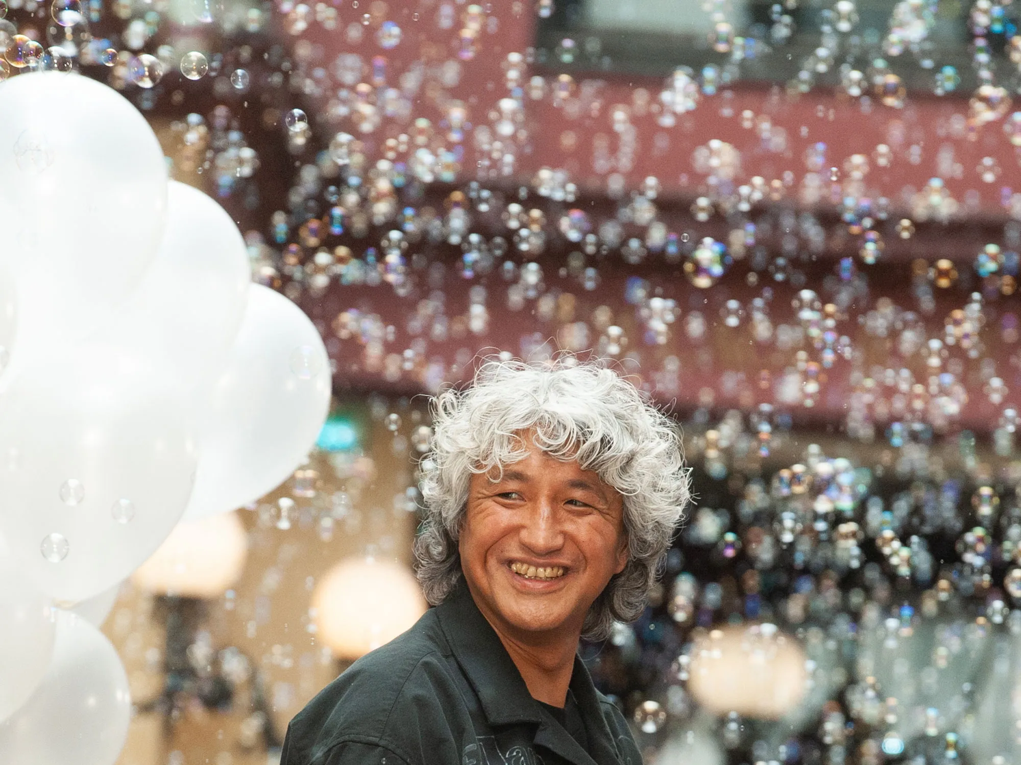 Yasuhiro Mihara smiles beneath a cascade of floating soap bubbles beside white balloons, capturing the playful and human side of the acclaimed Japanese designer during a candid moment