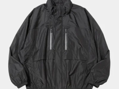 Lightweight technical wind jacket from meanswhile Spring/Summer 2026 in matte charcoal black, featuring a relaxed oversized silhouette, high-neck storm collar, reflective zip chest pockets, and softly crinkled performance nylon construction engineered for transitional weather and everyday urban mobility