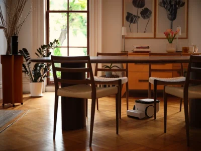 Matic robot vacuum cleaning beneath a wooden dining table inside a warm modern living space with hardwood floors, natural light, and minimalist interior decor