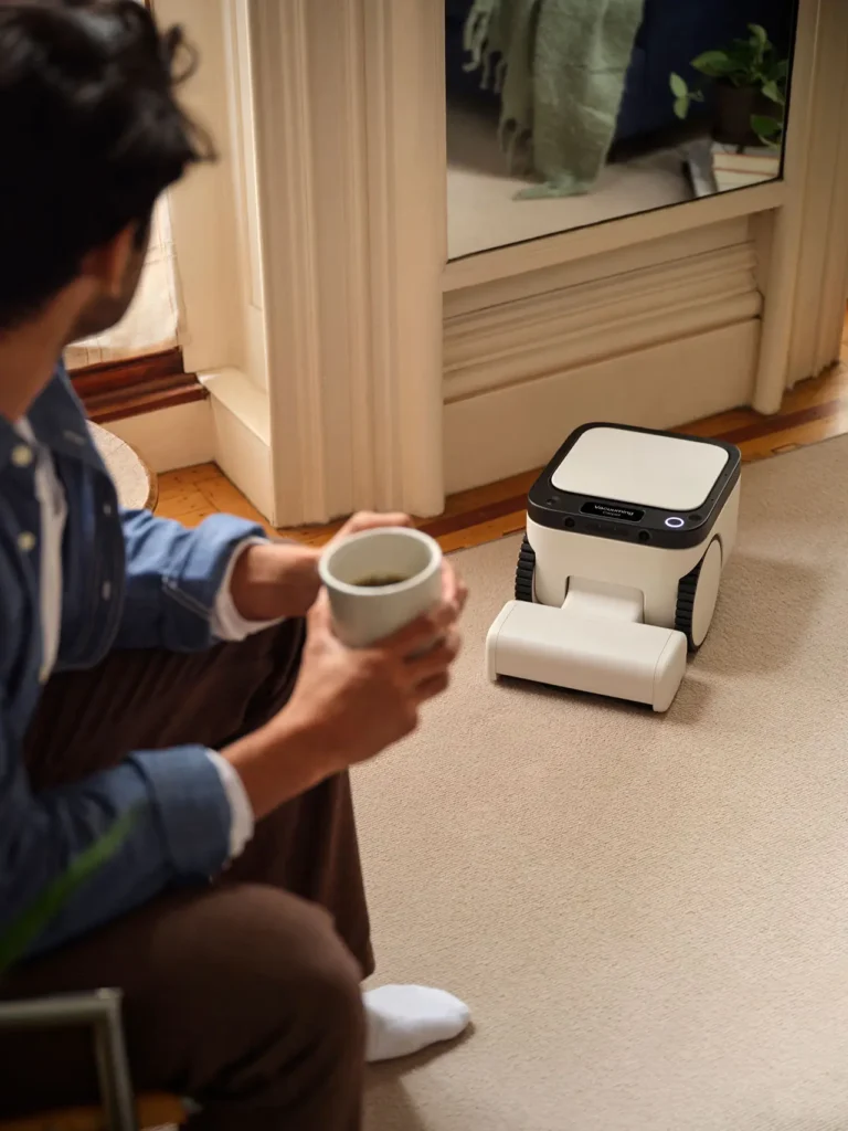 Person sitting indoors holding a mug while watching a Matic robot vacuum operate on a light-colored carpet inside a cozy modern home