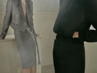 Two models pose within a softly fogged, industrial interior wearing looks from Alexander McQueen’s Autumn/Winter 2026 Pre-Collection under Seán McGirr. In the foreground, a model dressed in sculpted black tailoring wears oversized tinted sunglasses and dramatic shard-like statement earrings, her silhouette sharpened by ruched detailing and a cinched waist. Behind her, another model leans against a tiled structure in a cropped grey double-breasted jacket paired with a corset-laced pencil skirt and delicate embellished heels, blending rigid tailoring with sensual deconstruction. The muted lighting and misty atmosphere amplify the collection’s tension between structure, softness, and modern gothic elegance