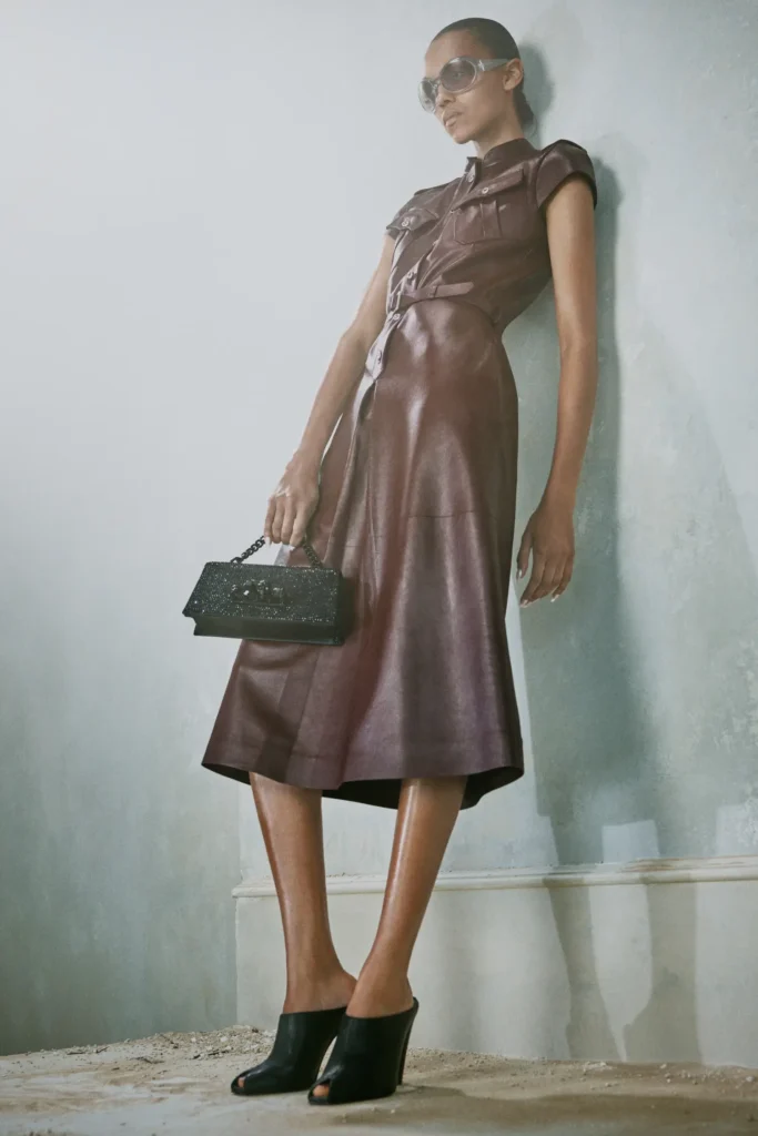 A model poses against a distressed interior wall wearing a sculpted oxblood leather dress from Alexander McQueen’s Autumn/Winter 2026 Pre-Collection by Seán McGirr. The sleeveless silhouette features sharp utility-inspired pockets, cinched paneling, and a fluid midi-length shape that merges rigidity with elegance. Oversized translucent sunglasses and black open-toe wedge heels amplify the futuristic edge, while a jeweled black top-handle bag introduces gothic glamour. The hazy lighting and weathered architectural backdrop reinforce the collection’s atmosphere of softened armor, modern sensuality, and urban sophistication