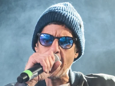 Close-up live performance portrait of a musician wearing a navy knit beanie and dark sunglasses while holding a microphone mid-vocal delivery. Cool-toned stage haze and bright backlighting create a cinematic concert atmosphere, while reflections in the lenses and the intimate framing emphasize the energy and immediacy of the performance moment