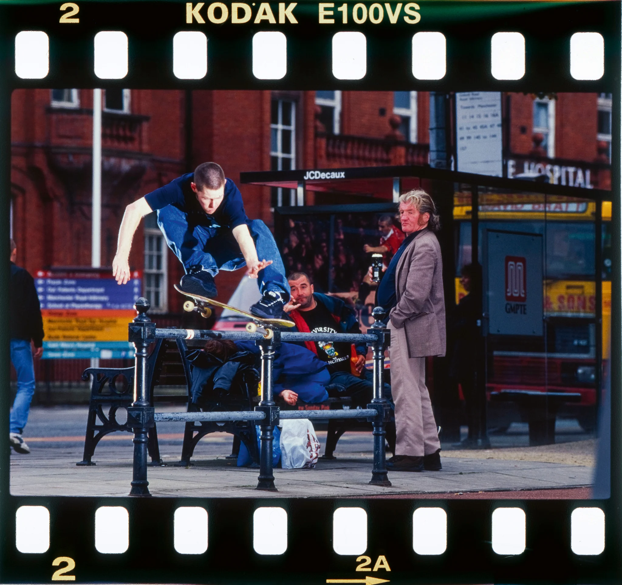 Archival skateboarding photograph presented as a mounted film slide, showing a skater mid-trick over black metal street barriers in a busy city setting while pedestrians and bystanders watch nearby—capturing the candid, documentary energy and public-space improvisation of early-2000s UK street skate culture