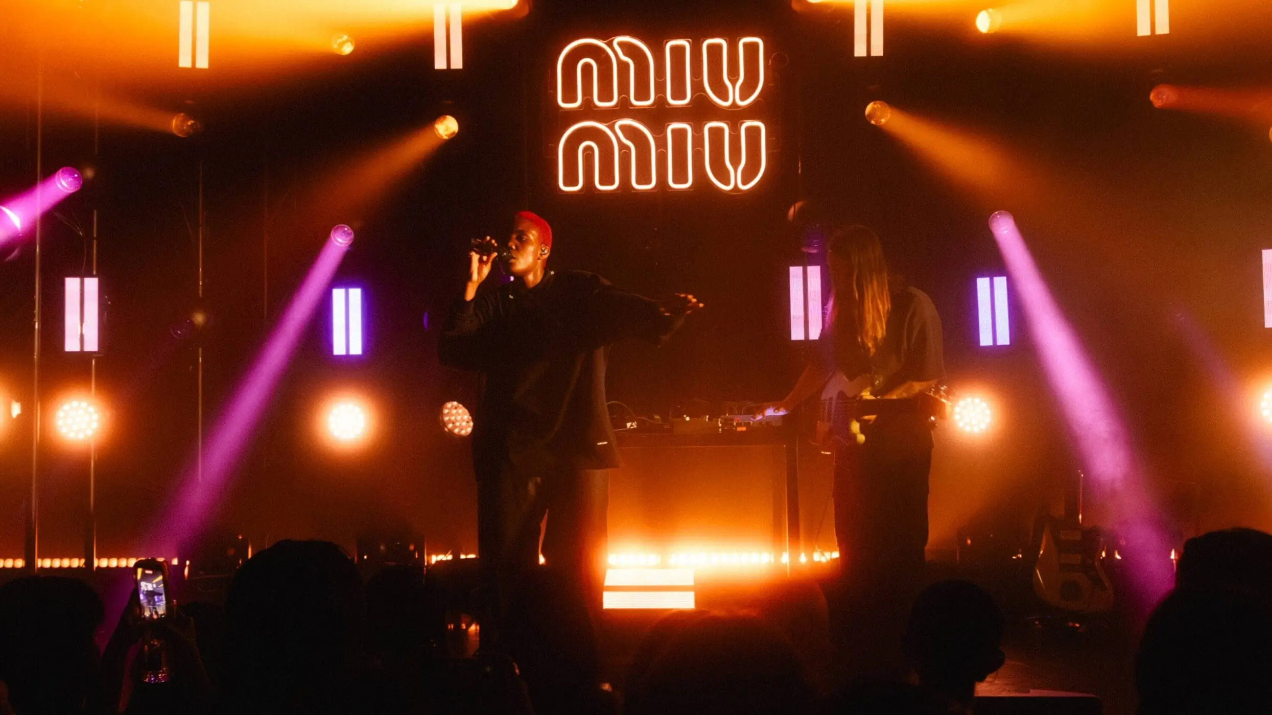 Live music performance beneath glowing Miu Miu signage with a vocalist and musician onstage surrounded by orange and purple lighting effects inside a dark concert venue