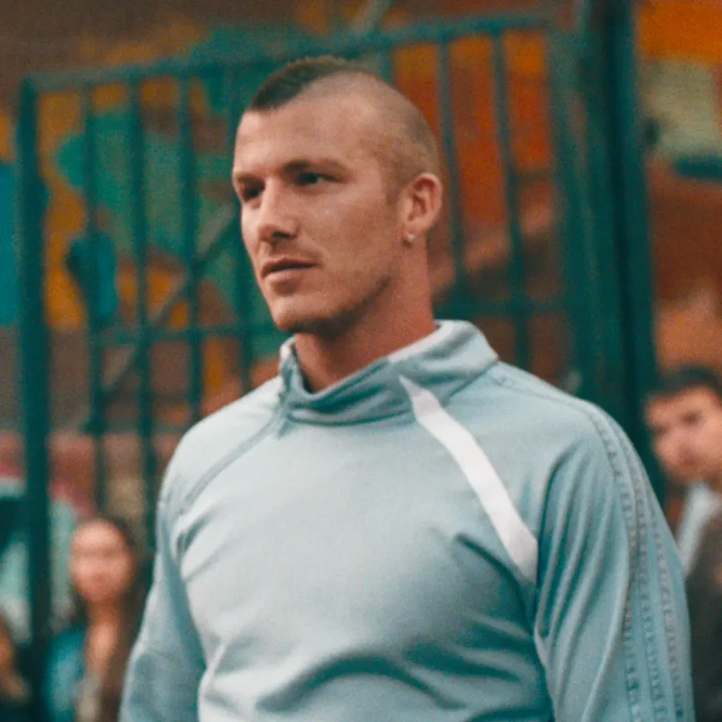 A man stands on an outdoor urban football court wearing a pale blue adidas track top with subtle tonal stripe detailing along the sleeves and white shoulder accents. His sharp mohawk hairstyle and small hoop earring give the look a distinctly early-2000s football aesthetic, while the softly blurred crowd and graffiti-covered fencing behind him reinforce the gritty neighborhood atmosphere. Warm cinematic grain and muted colors create a nostalgic portrait that channels classic street football culture and archival sportswear campaign imagery