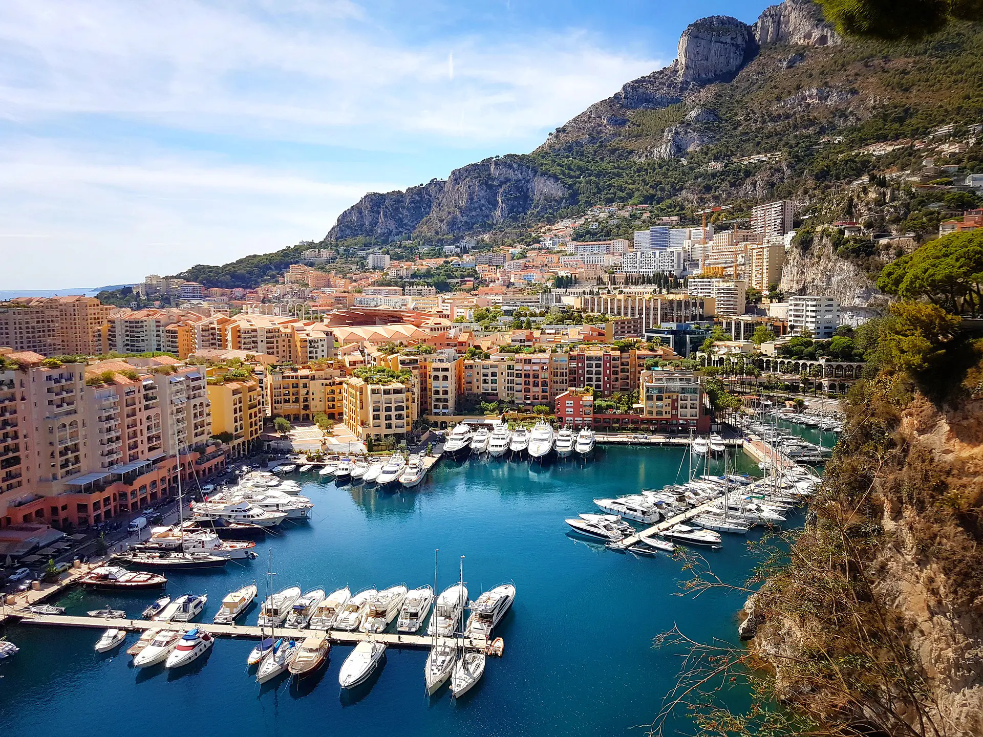 This sweeping view of Monaco highlights the principality’s signature mix of Mediterranean scenery and ultra-luxury lifestyle culture. Superyachts fill the vivid blue marina while warm-toned buildings rise dramatically against the surrounding cliffs and mountains