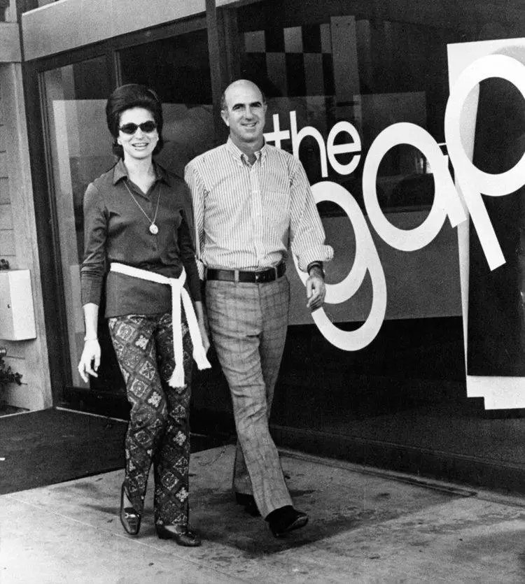A rare black-and-white archival photograph of Doris and Don Fisher standing outside an early Gap Inc. storefront during the company’s formative years. Doris wears patterned trousers, oversized sunglasses, and a sharply styled blouse that reflects the youthful energy and evolving style language of late-1960s and early-1970s American fashion, while Don appears beside her in tailored casualwear. The image captures the couple’s entrepreneurial optimism and the visual beginnings of a retail brand that would go on to reshape casual fashion culture across the United States and beyond