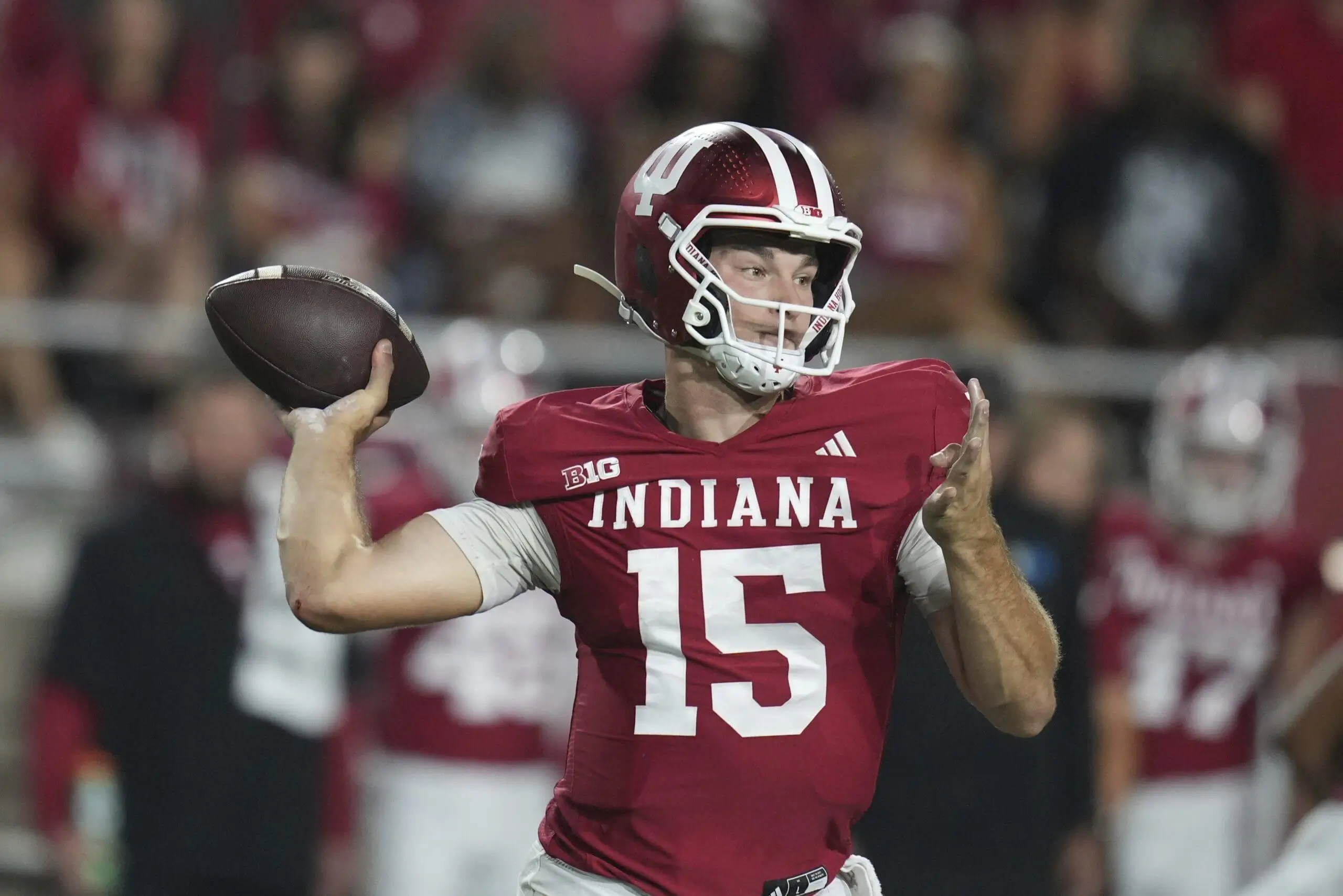 Quarterback in a red Indiana Hoosiers jersey (#15) and helmet cocks his arm to throw a football, looking right with a blurred crowd and teammates in the background