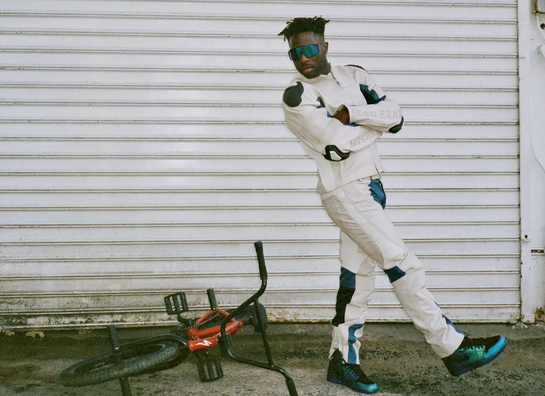 A stylized portrait of Nigel Sylvester posed against an industrial metal garage shutter while wearing a futuristic white-and-blue motocross-inspired riding suit. Oversized reflective visor sunglasses add a retro-tech aesthetic, while matching high-top sneakers coordinate with the blue paneling throughout the outfit. Beside him, a BMX bike lies overturned on the ground, reinforcing the raw street-riding atmosphere central to Sylvester’s identity. The grainy photographic texture and off-balance pose create a cinematic editorial mood that blends BMX culture, motorsport styling, and experimental streetwear imagery into a distinctly fashion-forward visual narrative