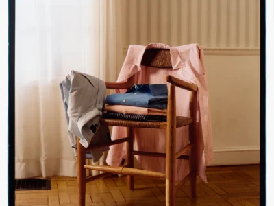 Alt text: Softly lit editorial still life of folded button-down shirts arranged on a woven wooden chair, with pastel pink and light blue shirts draped over the back and armrests. Set against sheer curtains and parquet flooring, the composition emphasizes timeless tailoring, natural textures, and understated haute