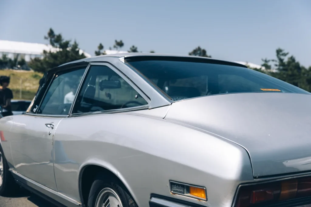 Rear-quarter detail of a silver Isuzu 117 Coupé highlighting the flowing fastback greenhouse, chrome window surrounds, sculpted rear fenders, and elegant classic GT proportion