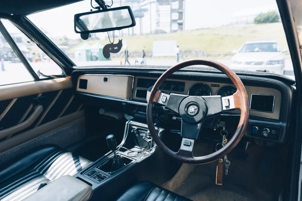 Driver-focused interior of an Isuzu 117 Coupé featuring a wood-rim steering wheel, analog gauges, manual transmission, and period-correct haute appointment