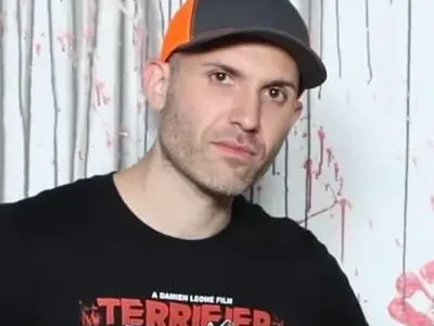 Damien Leone wearing a black Terrifier T-shirt and gray-and-orange cap while posing in front of a paint-splattered backdrop