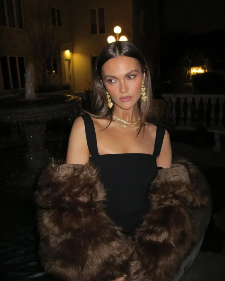 Nighttime portrait of a woman standing outdoors near a softly illuminated fountain and courtyard architecture. She wears a structured black square-neck dress styled with layered gold necklaces and long cascading gold earrings. A dark brown fur coat is draped loosely around her arms, adding texture and contrast. Her straight center-parted hair and understated makeup create a refined, editorial mood, while flash photography produces a glossy finish against the dimly lit background