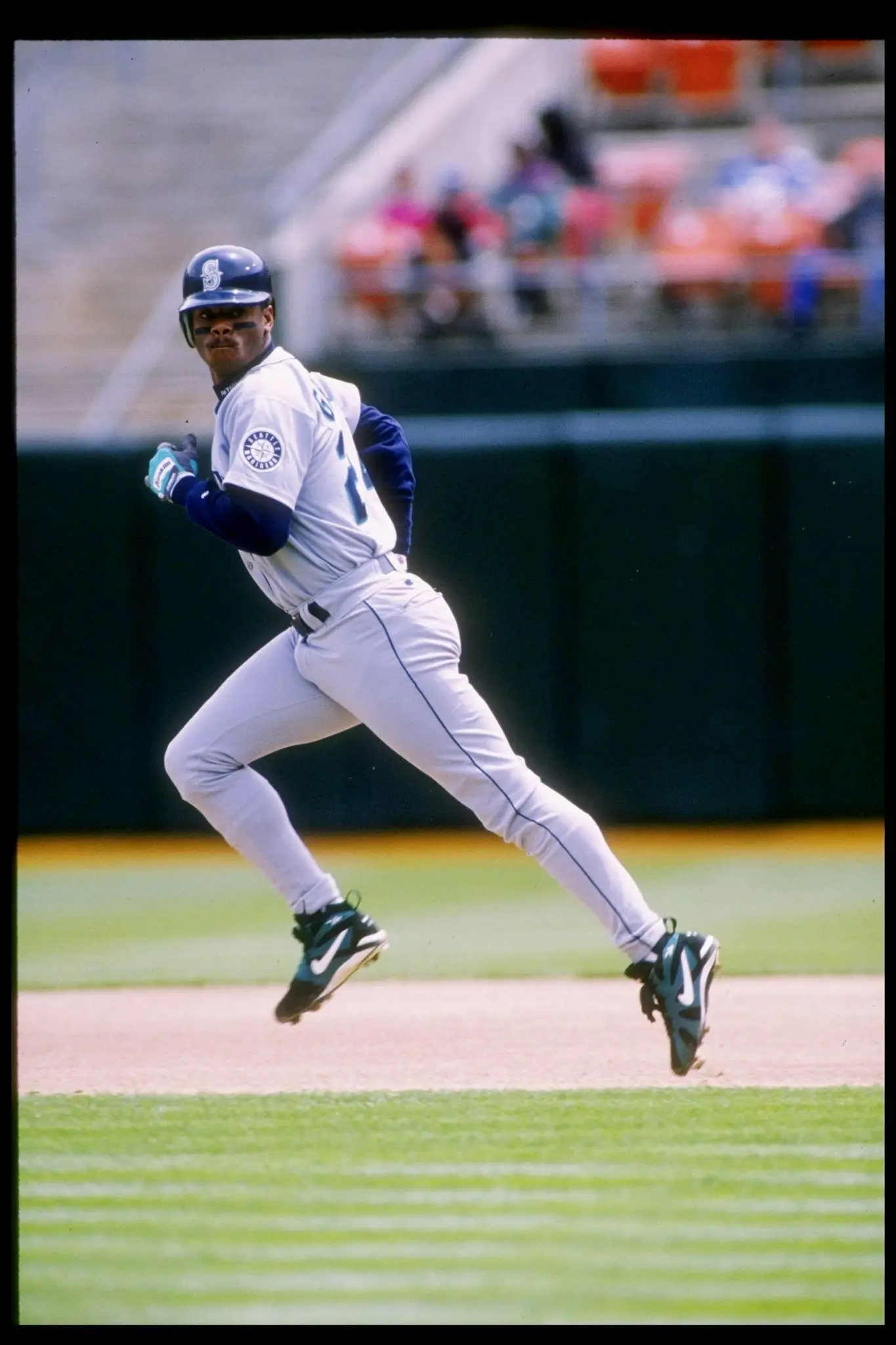 Vintage action photograph of baseball legend Ken Griffey Jr. sprinting across the field in a gray Seattle Mariners uniform during a game. Griffey is captured mid-stride with both feet off the ground, wearing black Nike cleats with teal accents and a navy batting helmet, while blurred stadium seating and spectators create a nostalgic 1990s sports atmosphere in the background