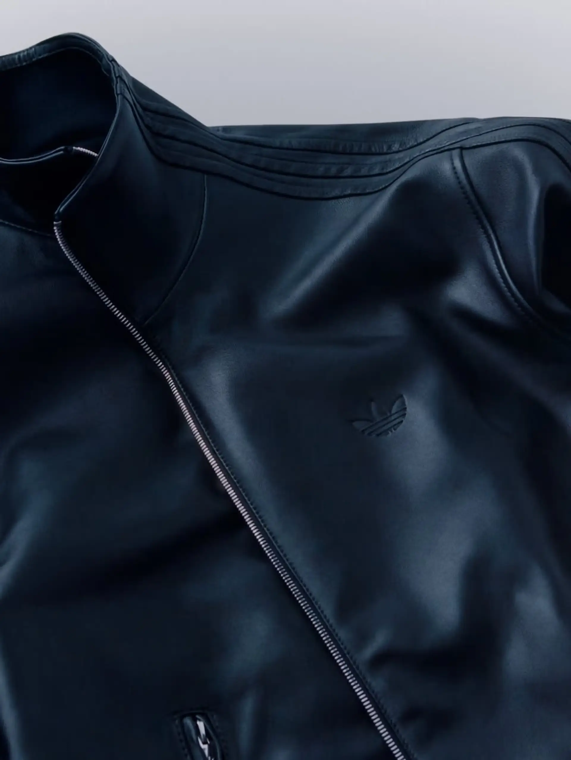 A detailed product close-up showcases a black Adidas leather track jacket crafted with a smooth matte finish and subtle tonal Trefoil branding at the chest. The elevated silhouette features a structured high collar, metallic zip closure, and refined stitched Three Stripes flowing across the shoulders, emphasizing the collection’s fusion of classic sportswear codes with minimalist luxury design