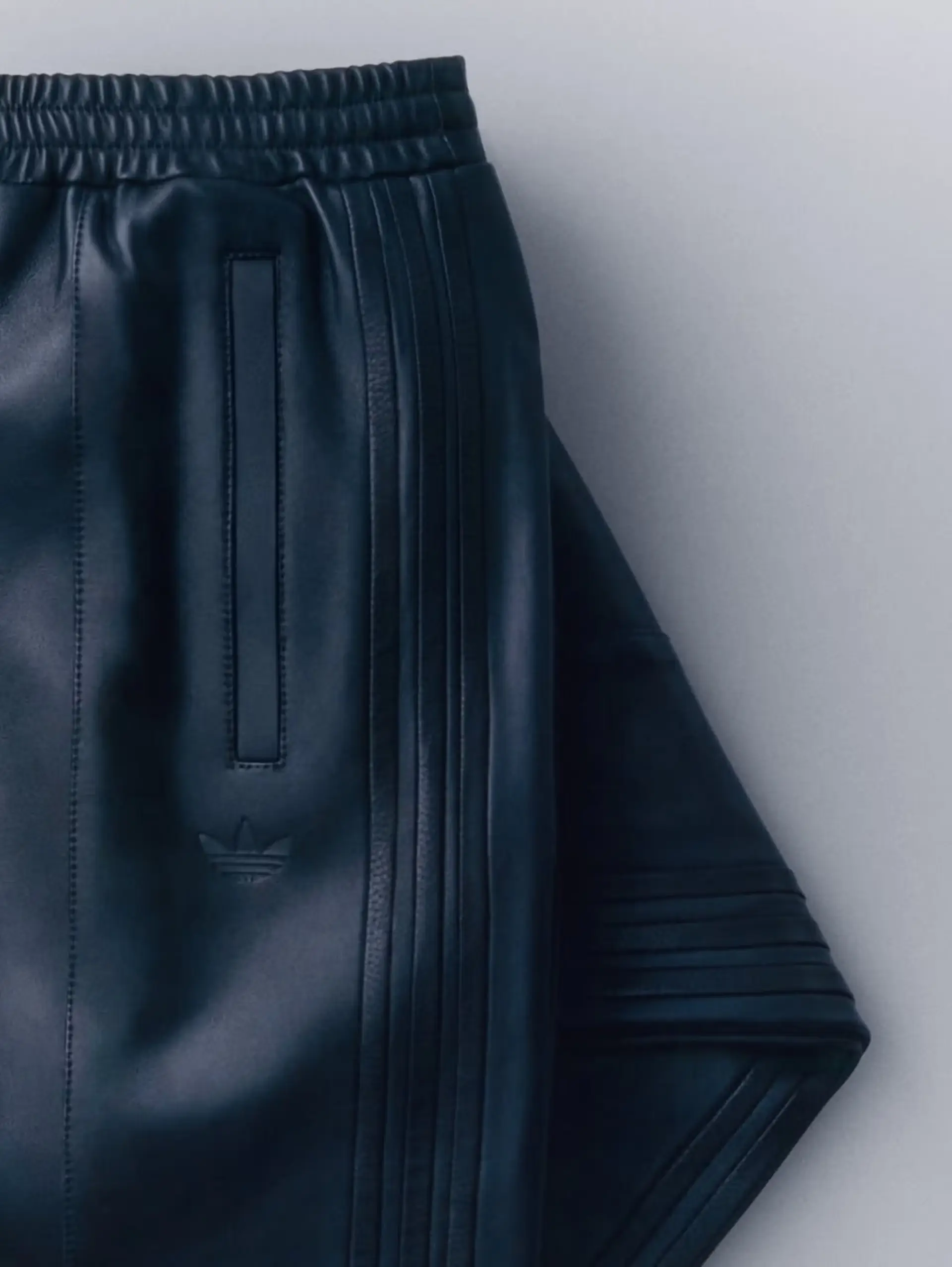 A close-up product shot highlights a pair of black Adidas leather track pants crafted with a smooth premium finish and tonal Three Stripes detailing running down the side seams. The minimalist design features an elastic waistband, sleek vertical pocket construction, and an embossed Trefoil logo, while the sculptural drape and monochromatic palette reinforce the collection’s elevated luxury-sportswear aesthetic