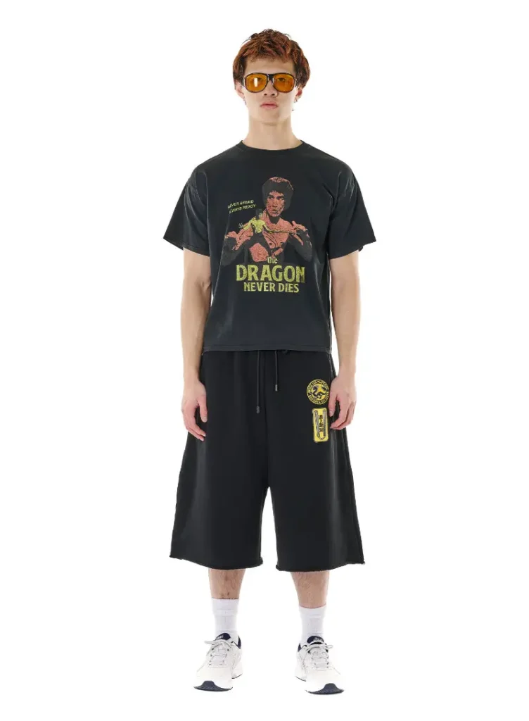 A model wears a graphic black tee from the LỰU ĐẠN x Bruce Lee collaboration featuring a distressed portrait of Bruce Lee above the phrase “The Dragon Never Dies.” Styled with oversized black athletic shorts accented by martial arts-inspired yellow patches, white socks, futuristic sneakers, and amber-tinted oversized sunglasses, the look blends gym-luxe streetwear with retro kung fu iconography. The relaxed silhouette and faded graphics channel Bruce Lee’s enduring cultural mythology through LỰU ĐẠN’s rebellious, contemporary menswear lens