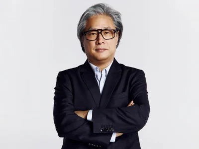 Portrait of filmmaker Park Chan-wook standing against a minimalist gray backdrop wearing a dark tailored suit, light blue shirt, and black glasses with arms crossed in a composed studio-style photograph
