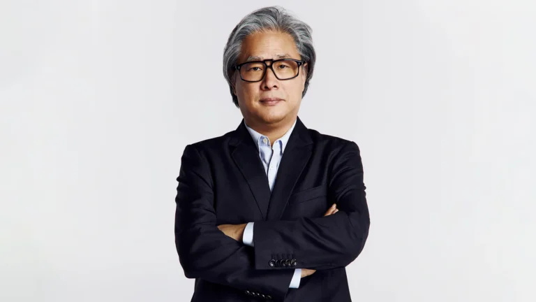 Portrait of filmmaker Park Chan-wook standing against a minimalist gray backdrop wearing a dark tailored suit, light blue shirt, and black glasses with arms crossed in a composed studio-style photograph