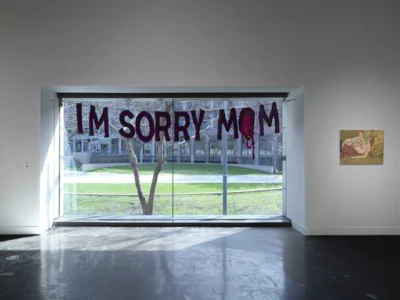 Installation view of Performing Conditions: Artistic Labor and Dependency as Form at the MIT List Visual Arts Center, featuring a large textile banner reading “I'M SORRY MOM” suspended across a floor-to-ceiling window overlooking the campus courtyard, alongside a figurative painting mounted on the gallery wall