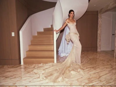 Kendall Jenner stands beside a sculptural curved staircase in a draped cream silk gown designed by Zac Posen for Gap Inc. Studio, featuring flowing Grecian folds, an elongated train, and soft marble-toned textures inspired by classical sculpture ahead of the Met Gala