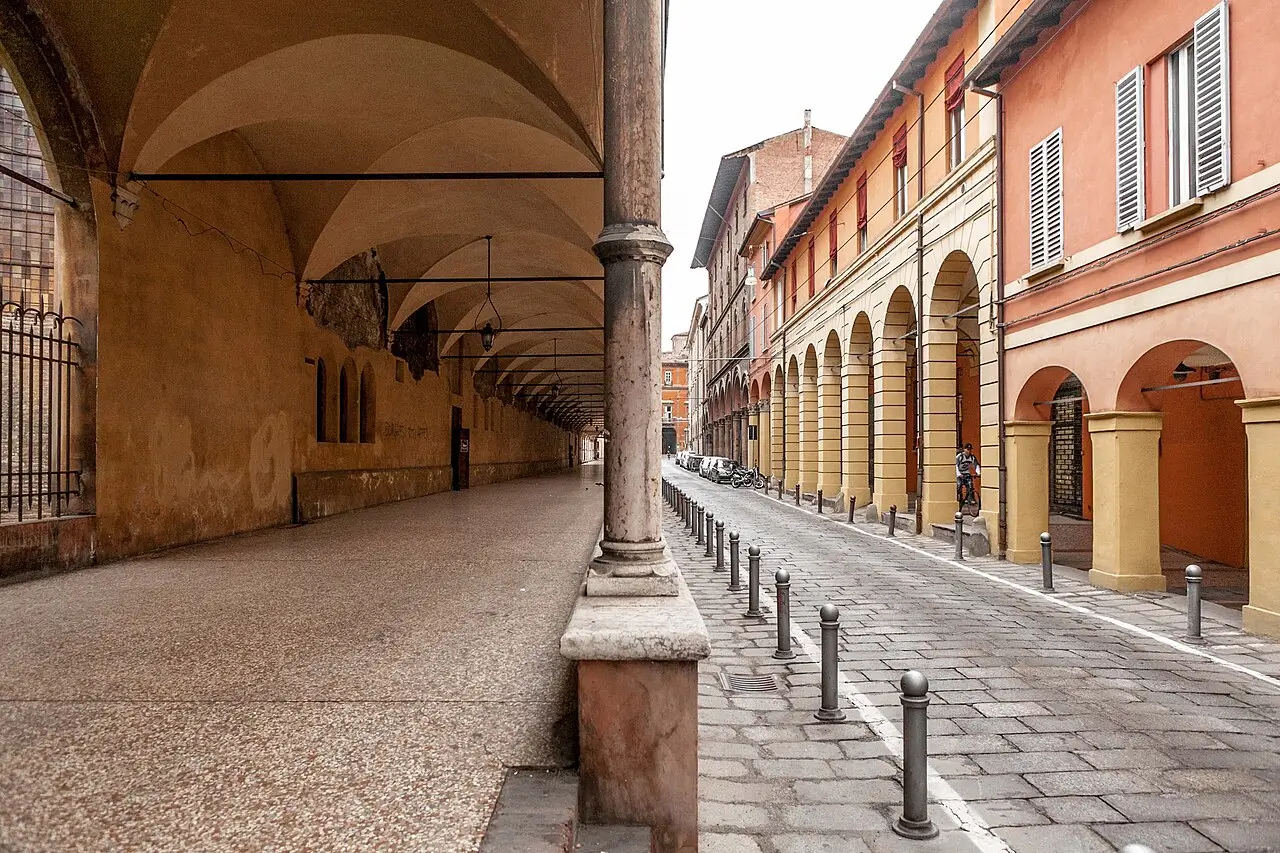 Covered arcade street in Bologna featuring classical Italian porticoes, stone columns, and pastel-toned historic architecture