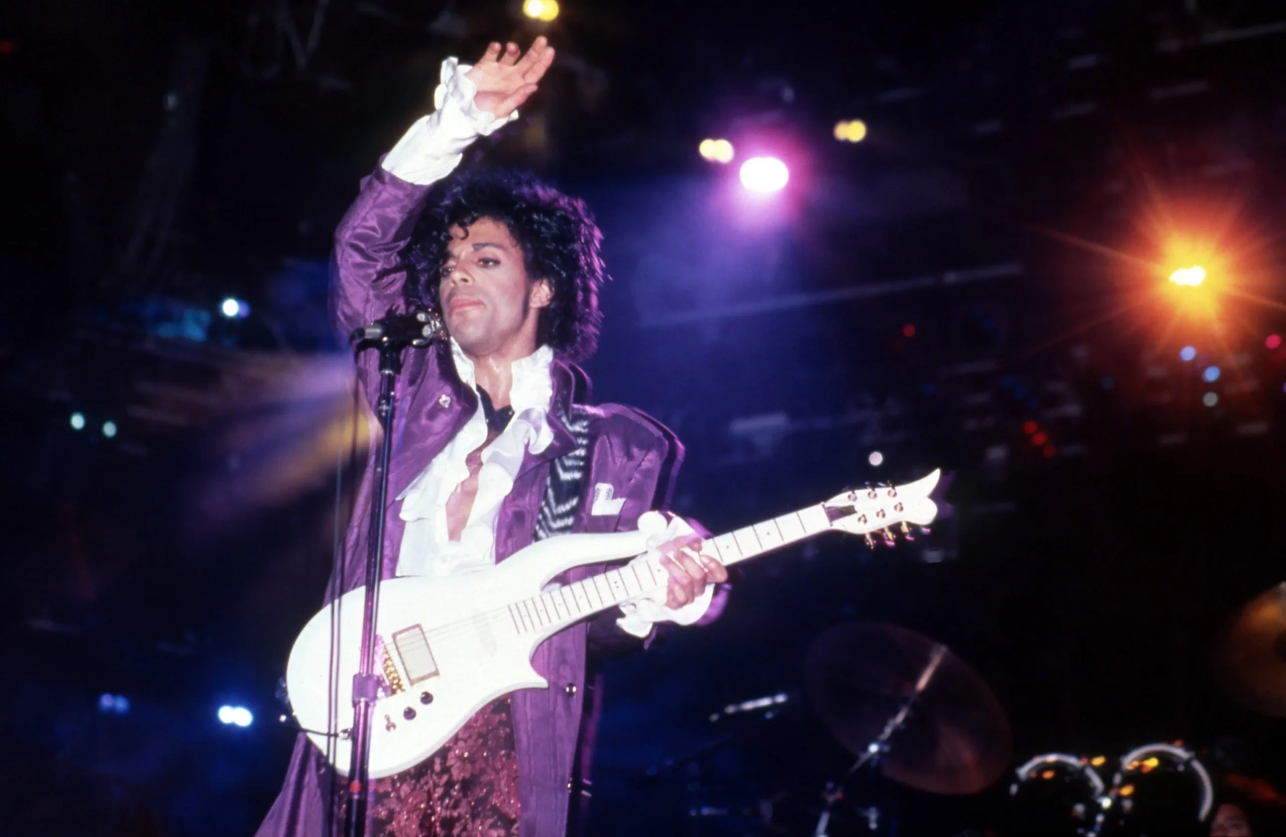 Archival concert photograph of Prince performing live in his iconic purple stagewear while holding a white electric guitar under vivid arena lights, capturing the theatrical energy and genre-defining charisma of the Purple Rain era