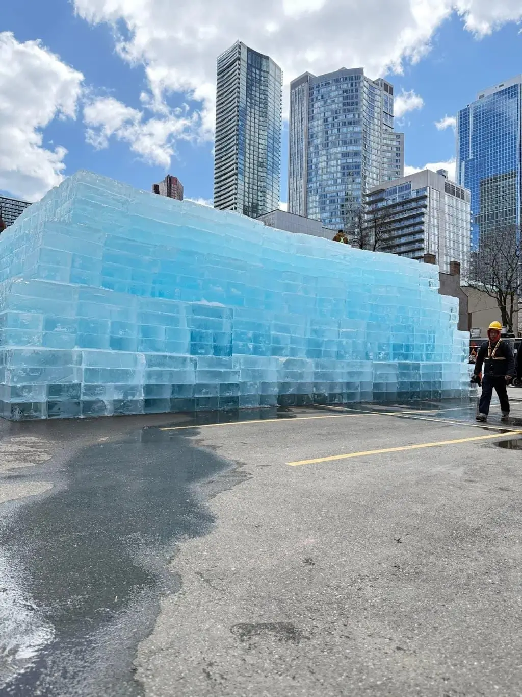 A massive translucent ice structure rises in the middle of a downtown Toronto parking lot, surrounded by towering glass skyscrapers under a bright blue sky scattered with clouds. Constructed from stacked frozen blocks tinted icy blue, the installation resembles a temporary frozen fortress slowly melting into the pavement below. Workers in safety gear stand nearby, emphasizing the sculpture’s monumental scale and industrial construction. The surreal contrast between the cold architectural mass and the surrounding urban skyline captures the theatrical rollout aesthetic of Drake’s Iceman era—turning public space into a cinematic spectacle rooted in mystery, anticipation, and controlled spectacle