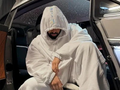 A moody portrait of Drake sitting inside a luxury Rolls-Royce with the door open, dressed in an oversized all-white hoodie and matching sweatpants. The hood is pulled low over his face, partially obscuring his eyes while emphasizing his beard and lowered posture. Holographic lettering runs vertically along the hood, adding a futuristic detail against the monochromatic fit. Above him, the car’s signature starlight headliner glows like a night sky, creating a cinematic atmosphere that reinforces the cold, isolated aesthetic associated with the Iceman era. The image blends luxury, introspection, and controlled mystique through muted lighting