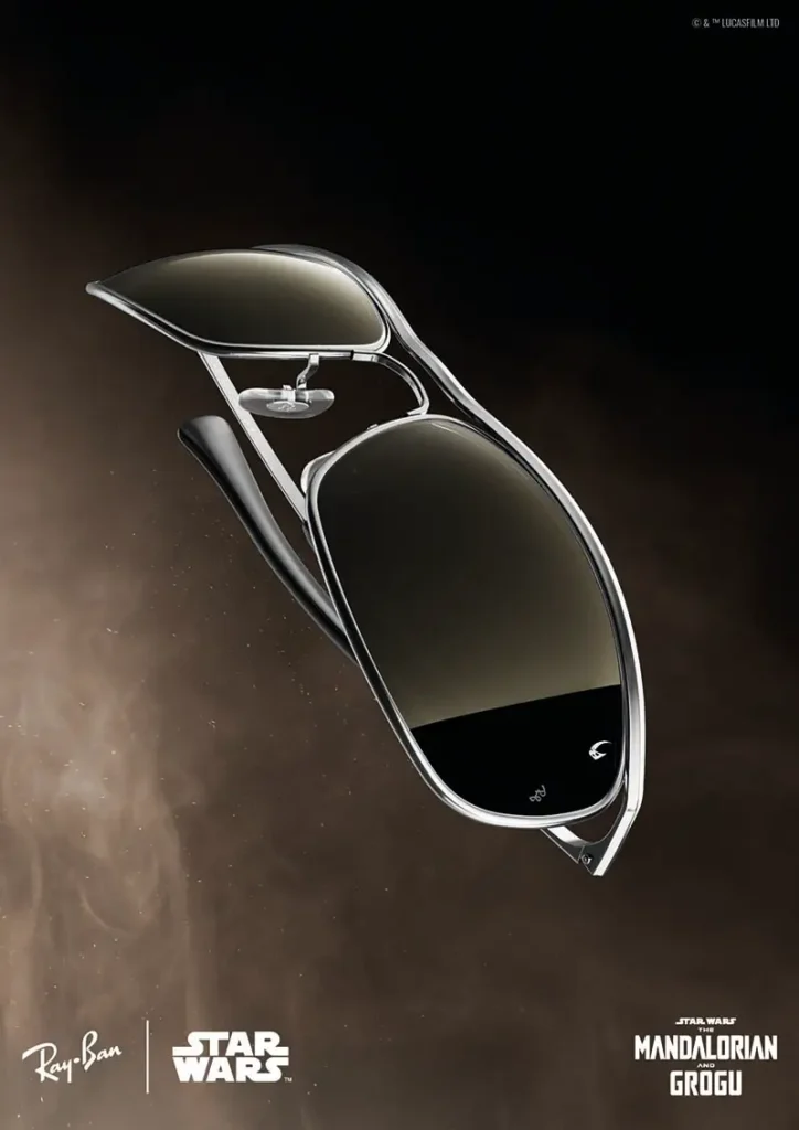 A sleek pair of futuristic Ray-Ban sunglasses floats against a dark, space-like backdrop in a cinematic promotional image tied to Star Wars: The Mandalorian and Grogu. Finished in reflective black lenses with ultra-thin silver metal framing, the eyewear channels a minimalist sci-fi aesthetic reminiscent of starship design and galactic technology. The composition emphasizes sharp aerodynamic curves and high-contrast lighting, while the collaborative branding from Ray-Ban, Star Wars, and Lucasfilm transforms the sunglasses into a luxury crossover object bridging fashion, cinema, and interstellar fantasy