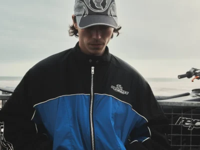 Model wearing a black-and-blue zip-up racing jacket and faded graphic cap stands against a metal bike trailer on a cloudy shoreline, with muted coastal lighting and motocross elements reinforcing the rugged, contemporary British moto-streetwear mood of the Belstaff x Represent collection