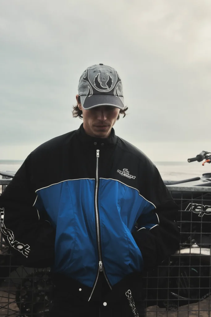 Model wearing a black-and-blue zip-up racing jacket and faded graphic cap stands against a metal bike trailer on a cloudy shoreline, with muted coastal lighting and motocross elements reinforcing the rugged, contemporary British moto-streetwear mood of the Belstaff x Represent collection