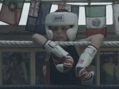 Young boxer wearing white protective headgear and Union Jack boxing gloves leans over the ropes inside a historic boxing ring. International flags hang overhead while framed photographs line the walls, capturing the traditional atmosphere and legacy of Repton Boxing Club in London’s East End