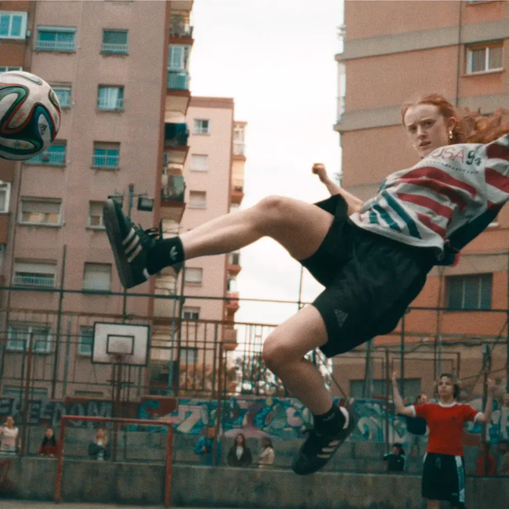 A young woman performs an airborne football kick on an outdoor urban court surrounded by tall apartment buildings and graffiti-covered fencing. She wears an oversized vintage USA World Cup graphic shirt paired with black adidas athletic shorts, striped socks, and classic black adidas sneakers as she twists midair toward a floating football. The dynamic motion, warm cinematic grain, and crowded neighborhood backdrop create an energetic street-football atmosphere rooted in community play, retro sportswear, and day city culture