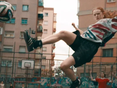 A young woman performs an airborne football kick on an outdoor urban court surrounded by tall apartment buildings and graffiti-covered fencing. She wears an oversized vintage USA World Cup graphic shirt paired with black adidas athletic shorts, striped socks, and classic black adidas sneakers as she twists midair toward a floating football. The dynamic motion, warm cinematic grain, and crowded neighborhood backdrop create an energetic street-football atmosphere rooted in community play, retro sportswear, and day city culture