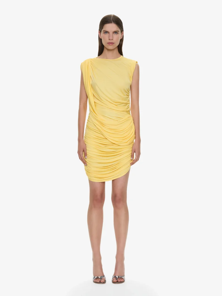 Model wearing a fitted pale yellow Christopher Esber mini dress with gathered draping and asymmetrical ruching throughout the silhouette, paired with metallic heels against a clean neutral studio backdrop