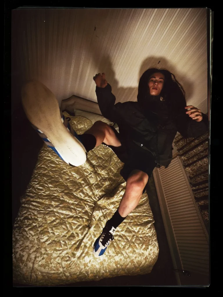 A dramatic overhead campaign shot from the Song For The Mute x adidas Originals “007” collection captures a model sprawled across a vintage bed while wearing a dark technical jacket and blue-accented retro adidas sneakers. The distorted perspective, shadow-heavy lighting, and grainy film texture create a dreamlike atmosphere that reinforces the collection’s themes of youthful disorientation, nostalgia, and imperfect memory