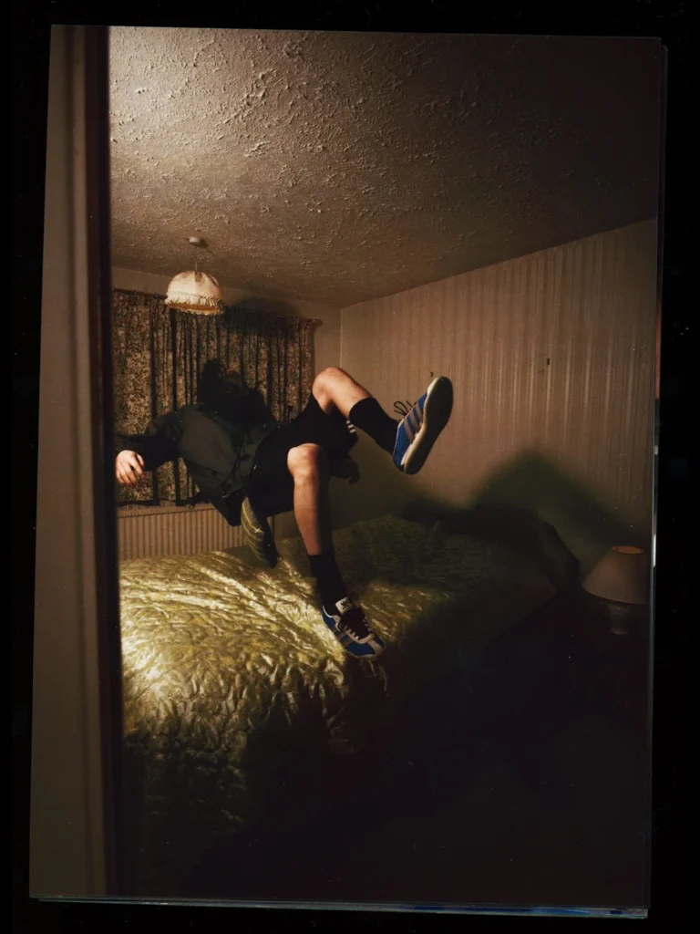 A moody campaign image from the Song For The Mute x adidas Originals “007” collection shows a model appearing to float above a vintage bed while wearing a dark technical jacket, black shorts, and blue-striped retro adidas sneakers. Shot with dim cinematic lighting and grainy film textures, the surreal bedroom scene amplifies the collection’s themes of distorted memory, adolescence, and emotionally charged nostalgia