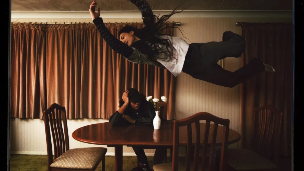 A surreal campaign image from the Song For The Mute x adidas Originals “007” collection depicts one model suspended midair above a dining table while another sits below with their head lowered, creating a dreamlike domestic scene. Warm cinematic lighting, vintage interiors, and floating movement reinforce the collaboration’s themes of nostalgia, adolescence, and emotionally worn-in storytellin