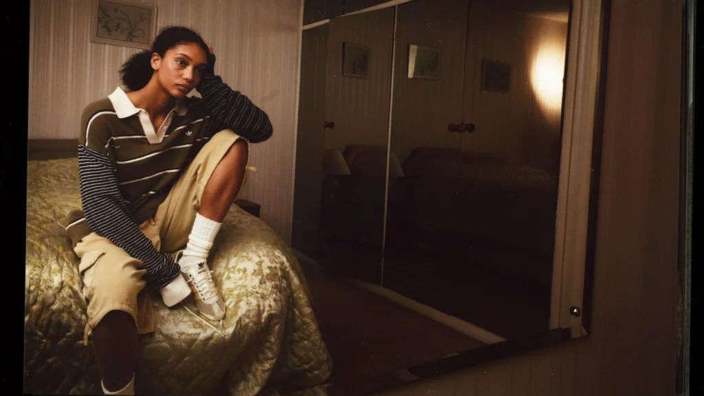 A campaign portrait from the Song For The Mute x adidas Originals “007” collection shows a model seated on a bed inside a softly lit vintage bedroom, styled in layered striped rugby-inspired separates, oversized shorts, and retro adidas sneakers. The warm cinematic tones and intimate domestic setting reinforce the collection’s themes of youthful nostalgia, quiet reflection, and imperfect everyday styling