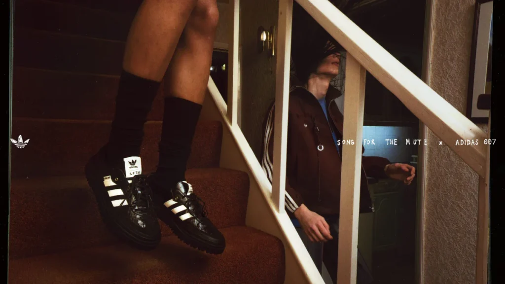 A close-up campaign image from the Song For The Mute x adidas Originals “007” collection spotlights the black Samba LX Freizeit silhouette with glossy leather construction, cream Three Stripes, co-branded tongue tags, and chunky lugged soles. Set against a dimly lit staircase interior, the cinematic composition blends distorted movement and nostalgic domestic imagery to emphasize the collaboration’s worn-in, emotionally charged aesthetic