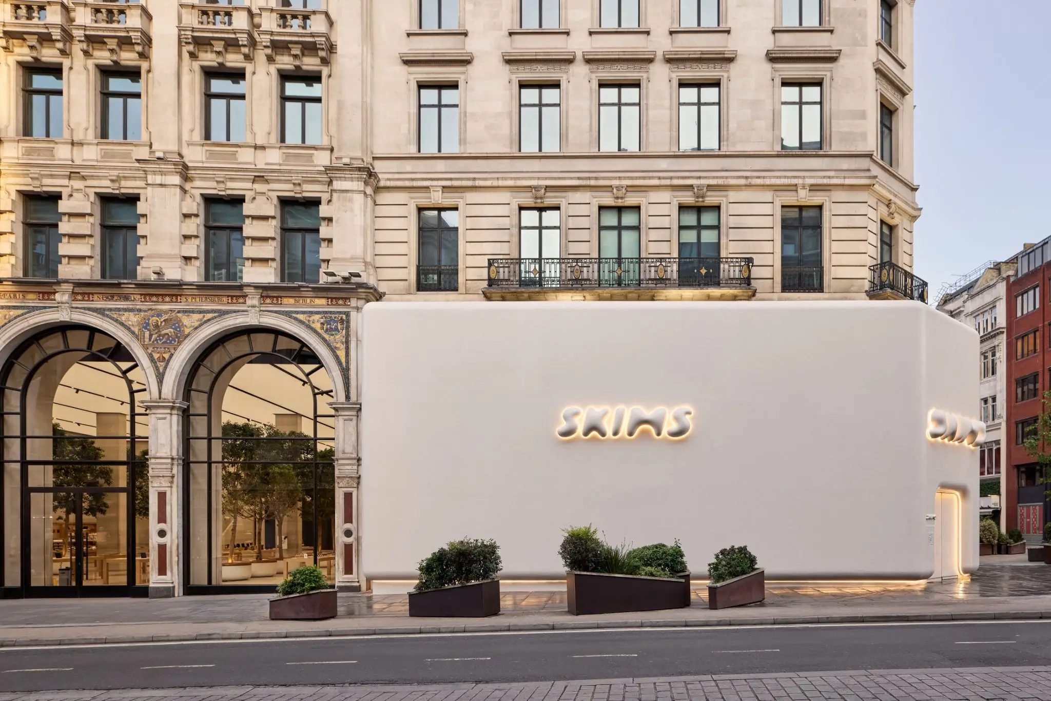 Exterior view of a SKIMS storefront installation set against a historic city building façade. A smooth, oversized cream-colored temporary structure wraps the street-level frontage, featuring softly illuminated SKIMS signage and a rounded entrance opening. Minimal landscaping planters line the sidewalk, creating a stark contrast between the contemporary, sculptural retail design and the ornate classical architecture surrounding it