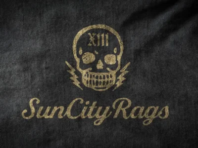 Close-up of a black Sun City Rags t-shirt featuring a distressed gold skull logo with “XIII” on the forehead, lightning bolt details, and the brand’s signature script typography printed beneath in a faded vintage style